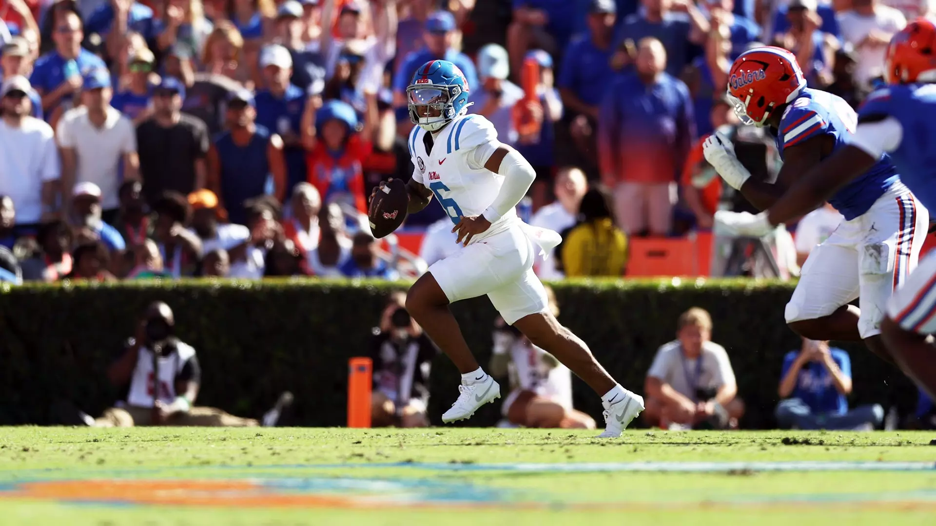 A football player in a white uniform runs with the ball as defenders in blue and red gear pursue him during a game in front of a large crowd.